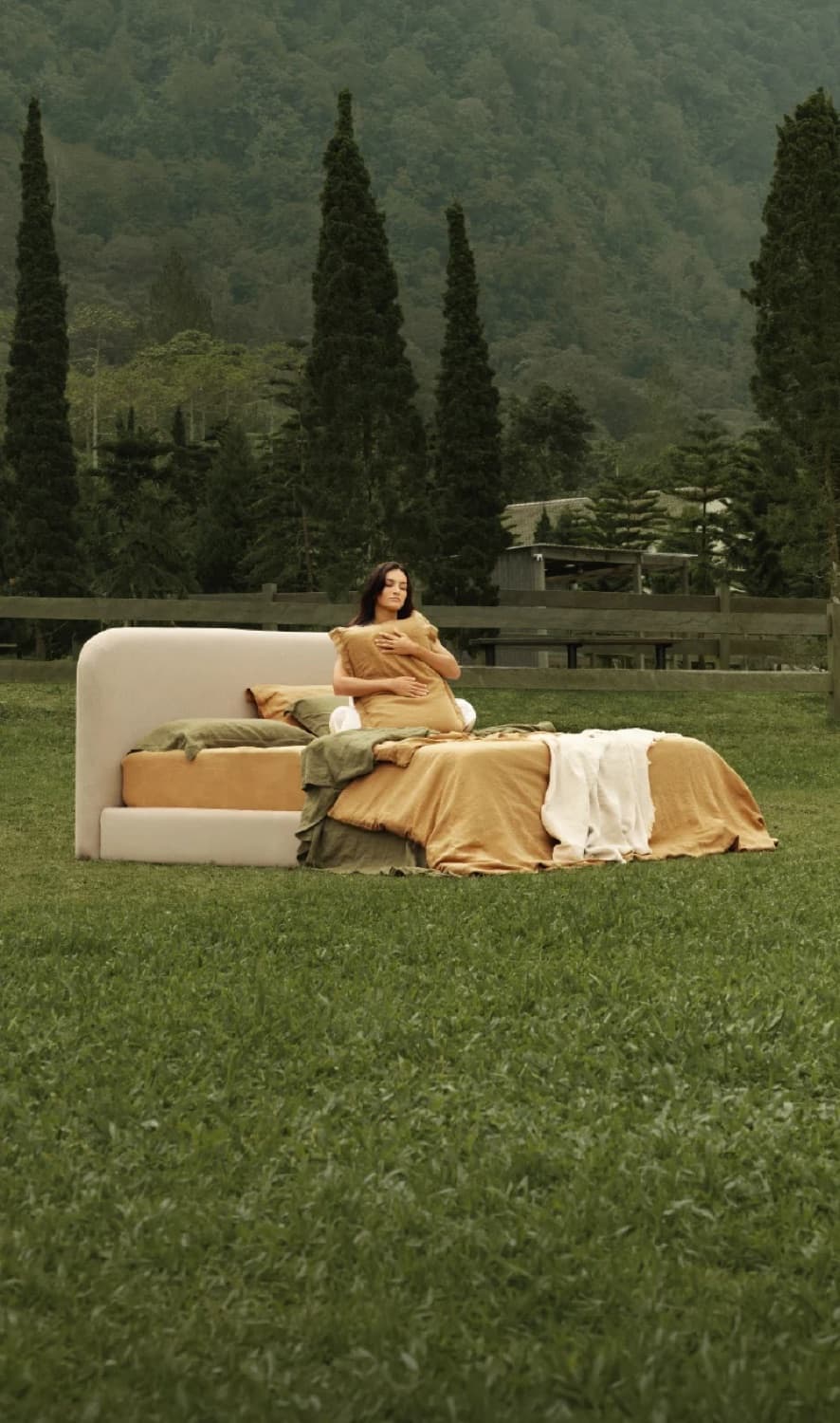 A woman sitting up in a Heveya bed made up on an open lawn, holding a pillow, mountains and cypress trees behind her.