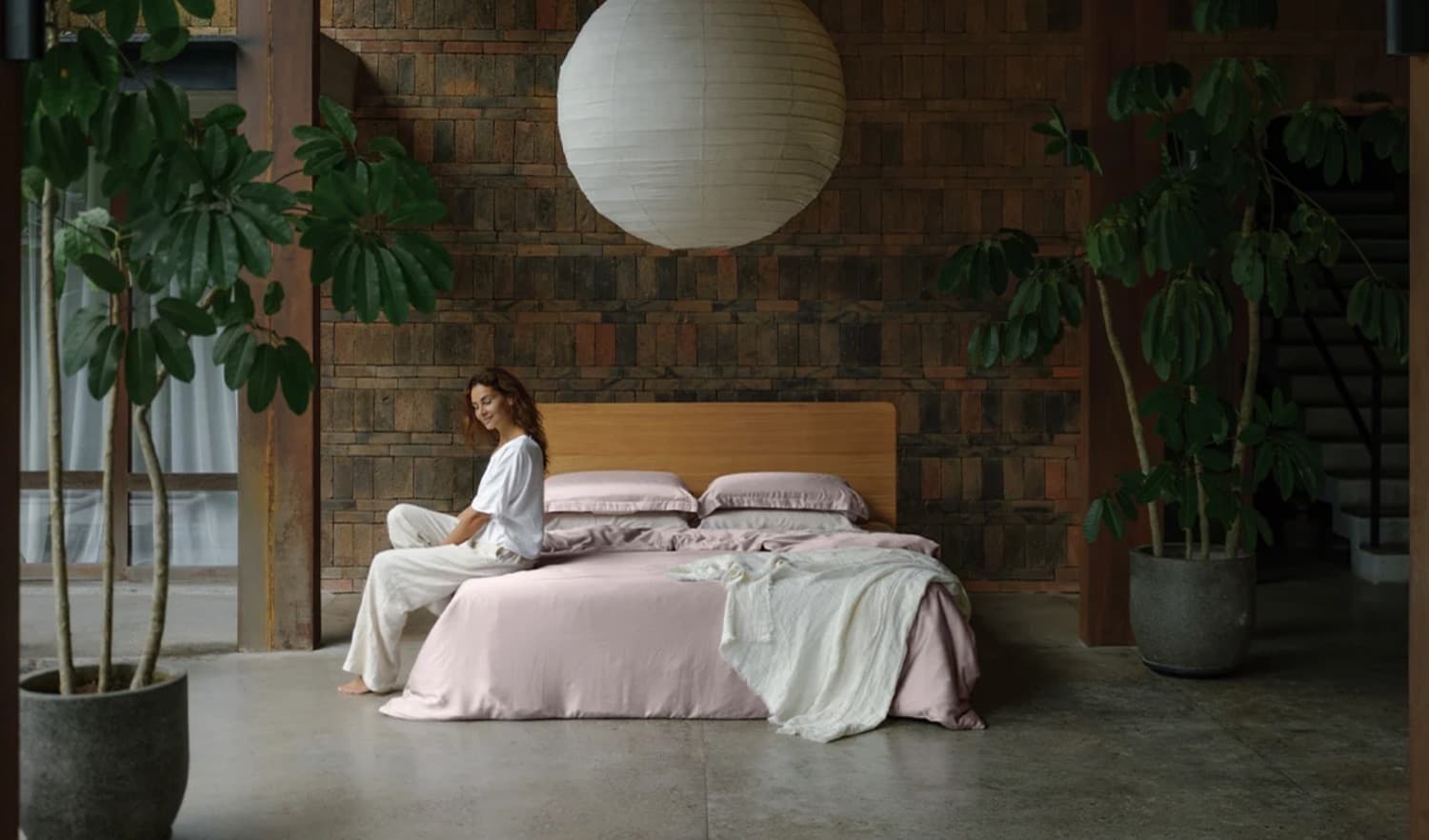 A woman sitting on the edge of a bed dressed in dusty pink linen, in a brick-walled room with tall plants and a paper lantern overhead.