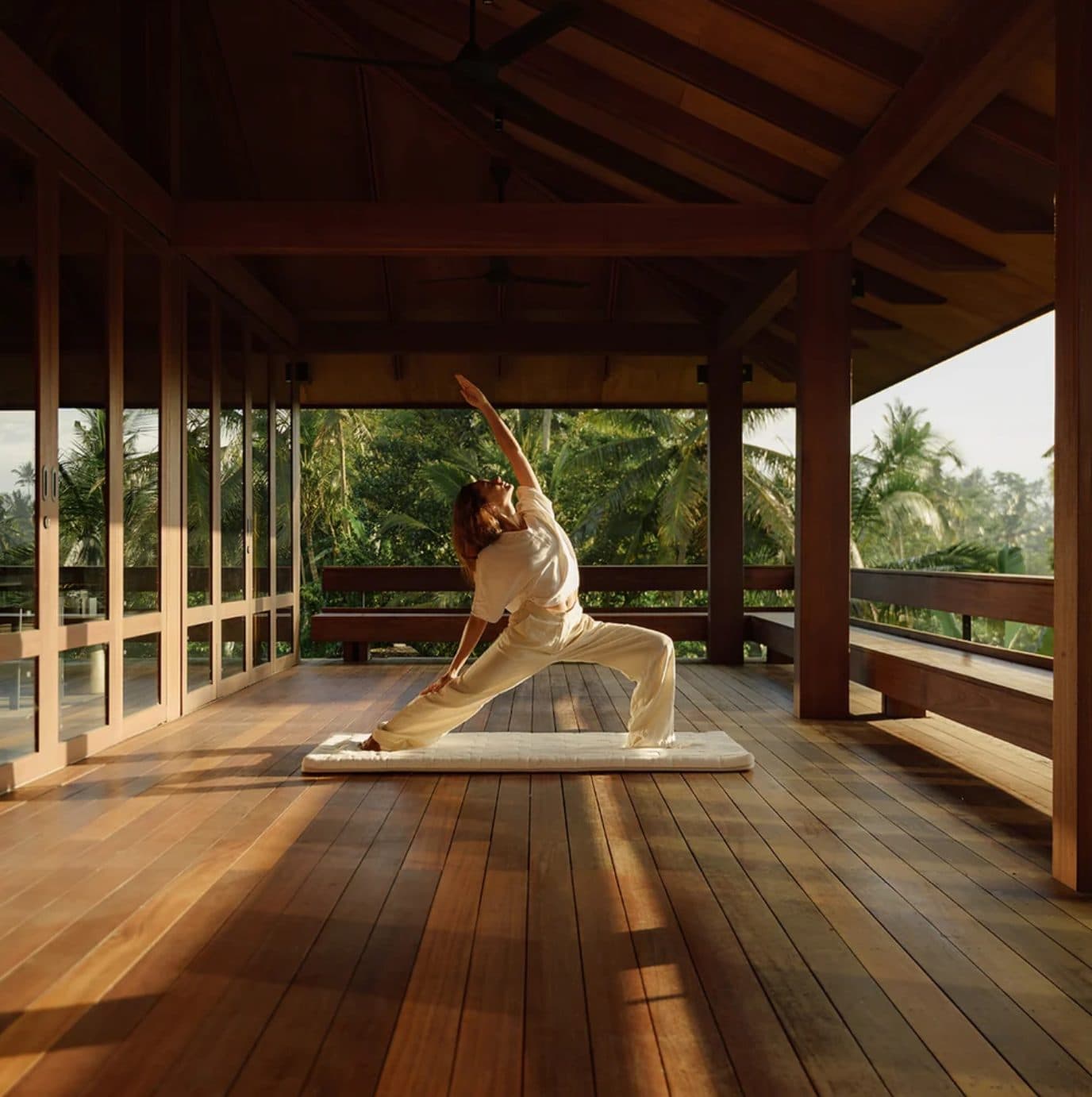 A woman stretching on a Heveya mat on the wooden floor of an open pavilion, palms beyond the railing.